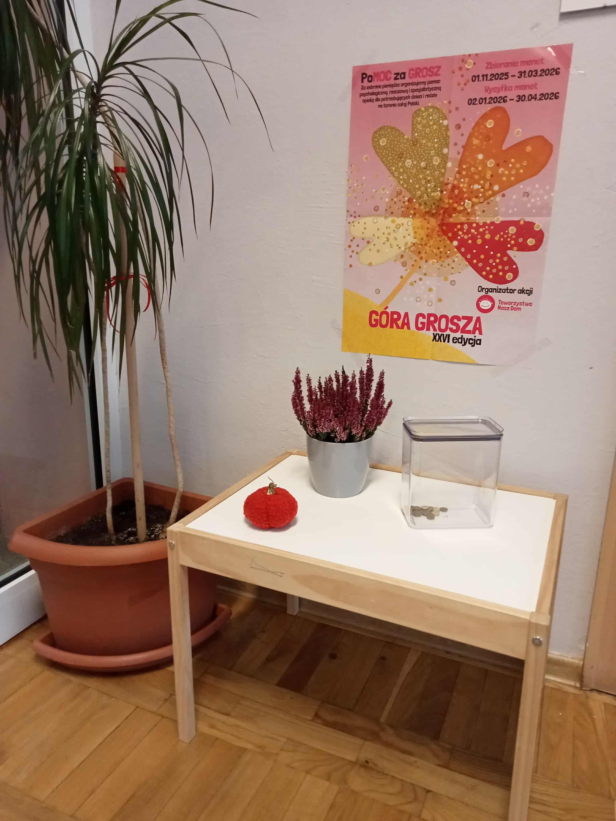 Zdjęcie miejsca zbiórki monet. Mały stoliczek na którym znajduje się puszka na monety. Nad stolikiem powieszony plakat informacyjny na temat zbiórki.