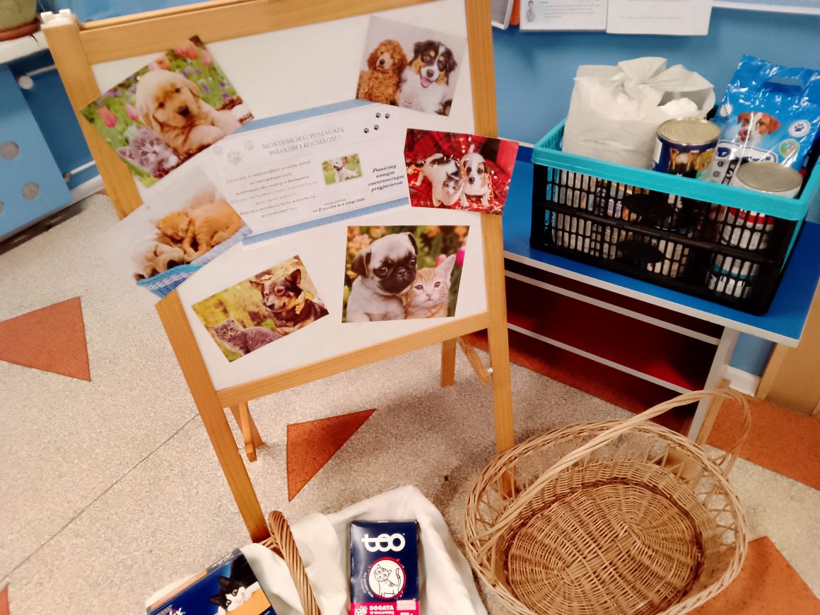 Zdjęcie miejsca zbiórki. Tablica z ilustracjami psów i kotów. Obok tablicy kosze na karmę.