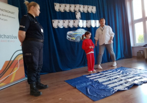 Na zdjęciu pani policjantka pokazuje dzieciom prawidłowy sposób przechodzenia przez przejście dla pieszych.