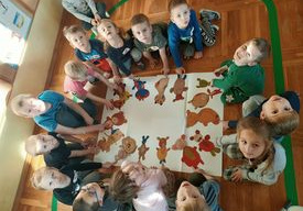 Dzieci siedzą na podłodze i tworzą kompozycję ze swoich papierowych misiów.